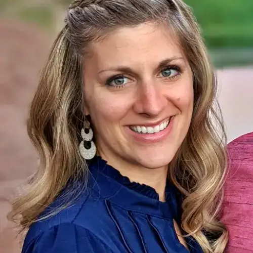 Smiling woman in a blue blouse with wavy blonde hair and dangling earrings, outdoors.