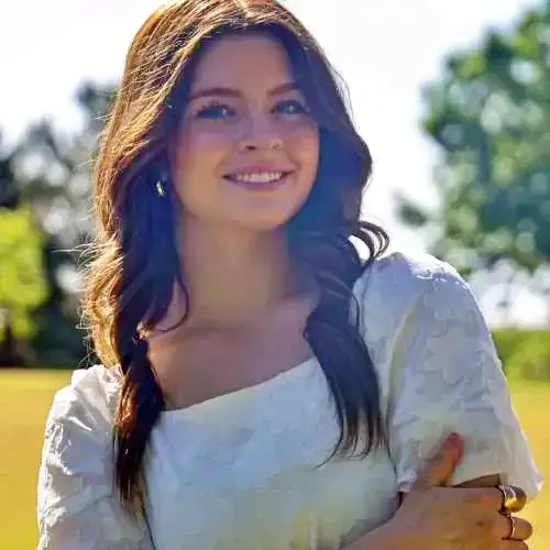 Smiling woman in a white dress standing outdoors in sunlight with trees behind her