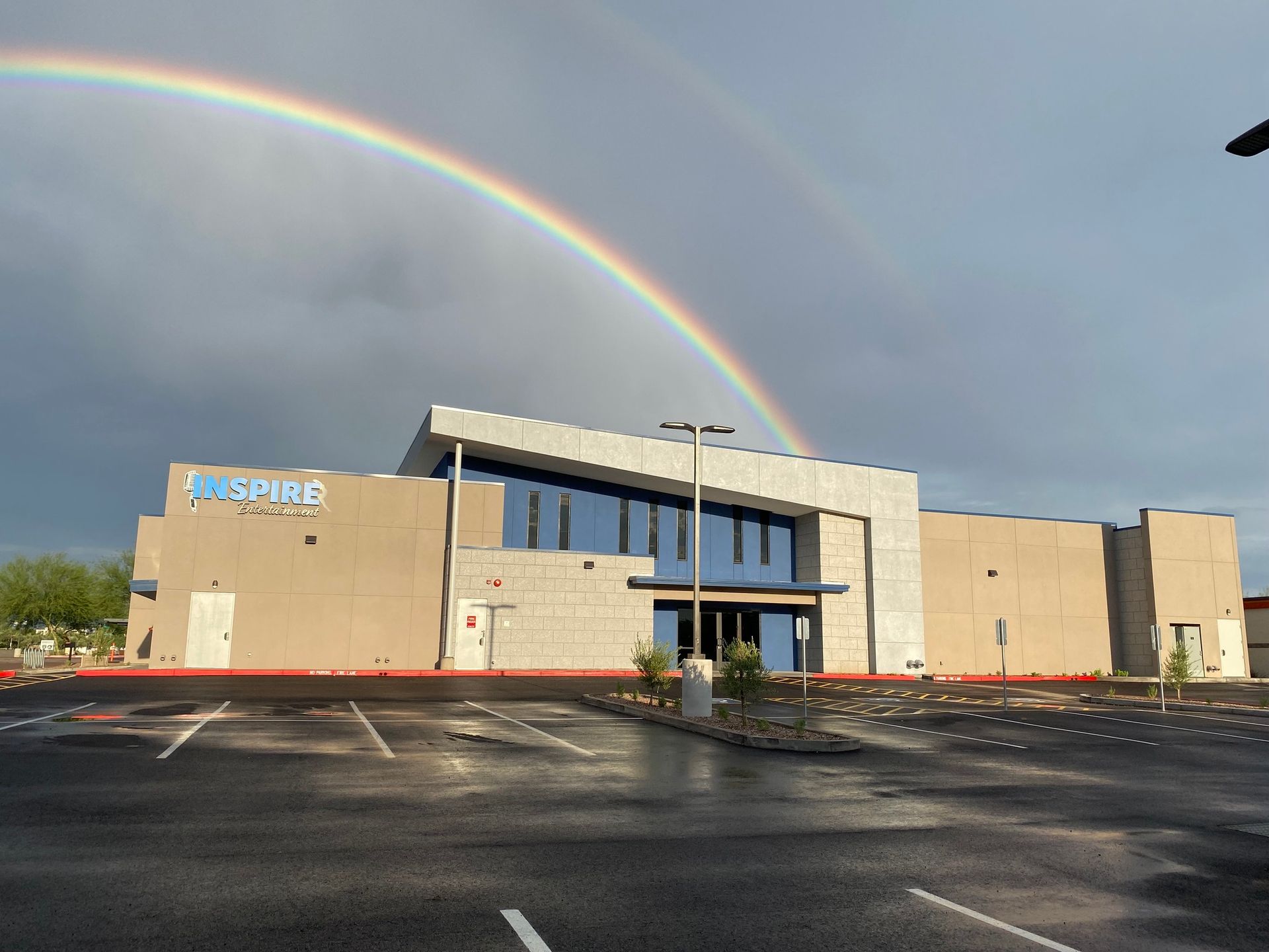 Modern beige building with a rainbow overhead and wet parking lot under cloudy sky