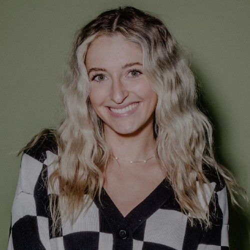 Smiling person with wavy blonde hair wearing a black-and-white checkered top against a green background