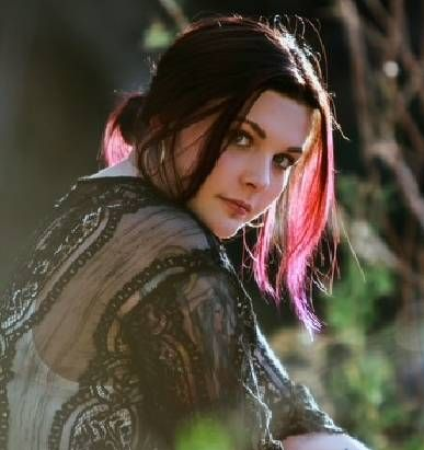 Person with pink-tinted hair in a black lace top looking over shoulder outdoors