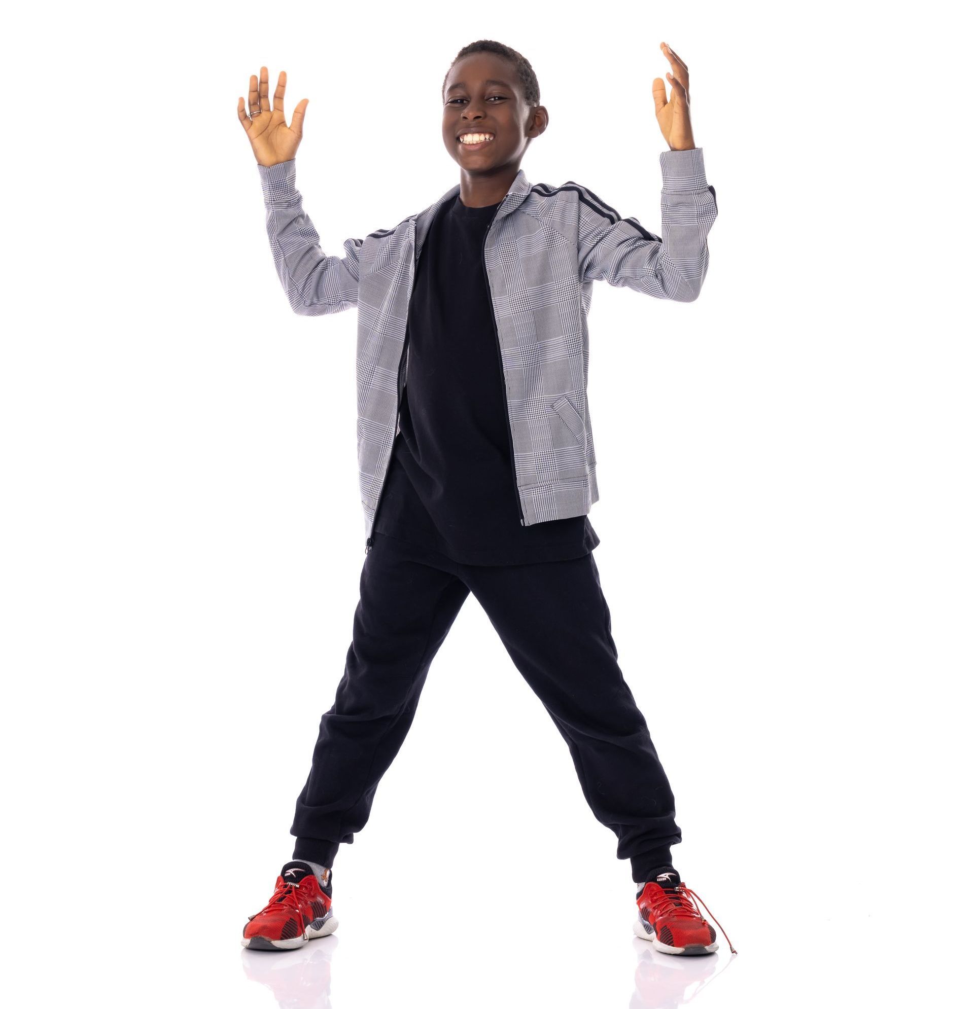 Person standing with raised hands, wearing a black outfit, patterned jacket, and red sneakers on a white background