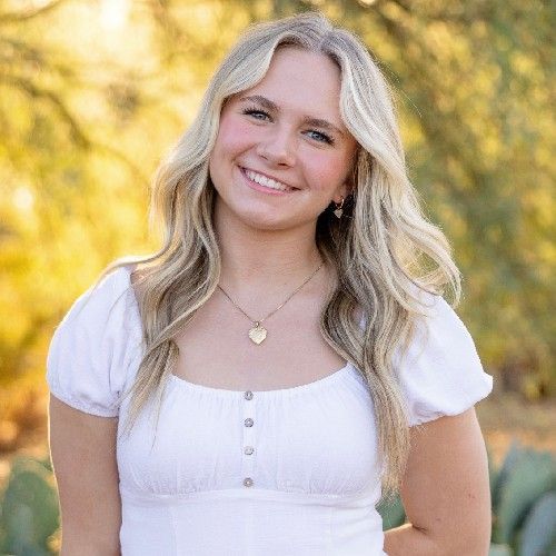 Smiling blonde woman in a white top outdoors with warm, sunlit trees behind her