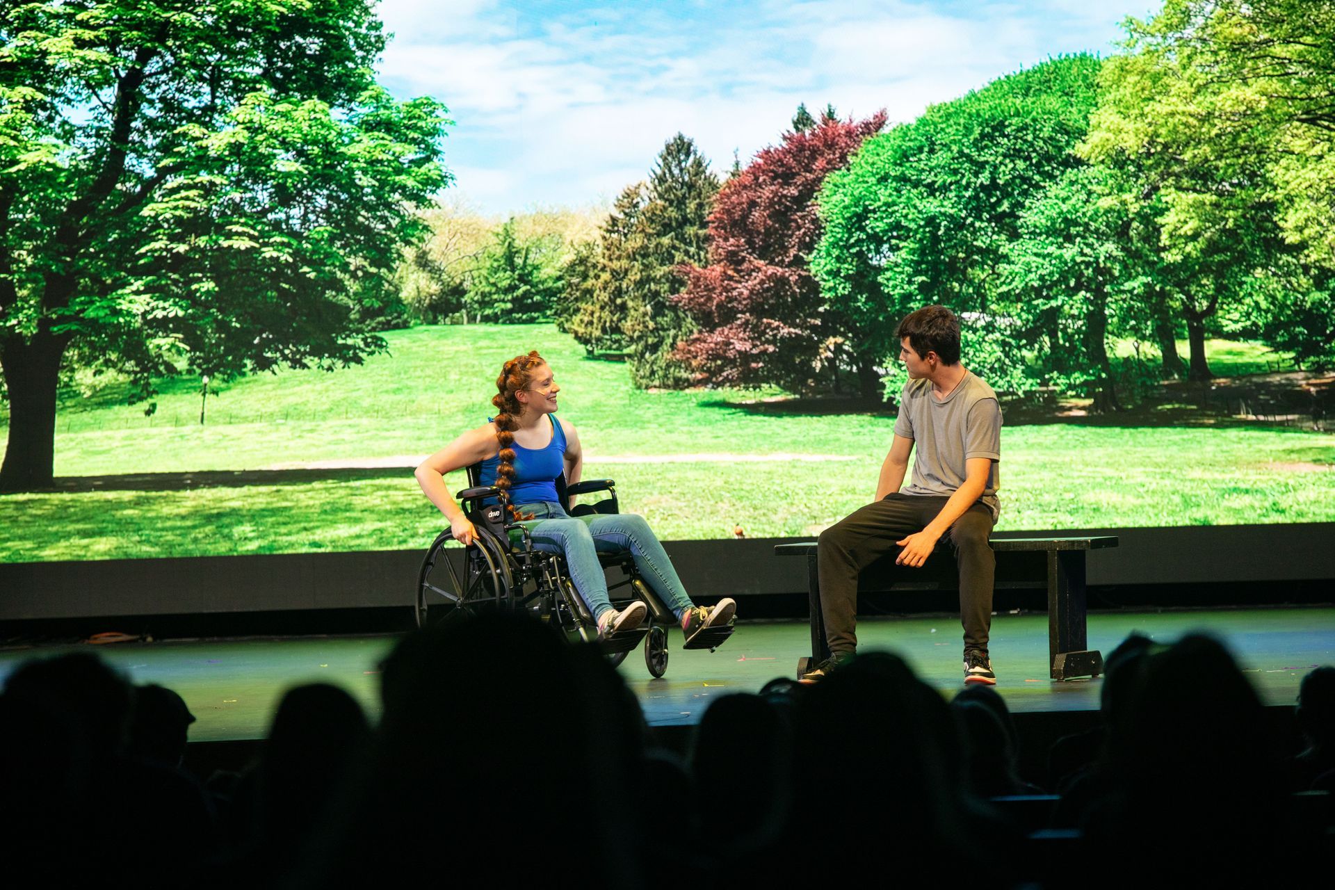 Two people seated onstage in front of a bright green park backdrop, speaking during a presentation.