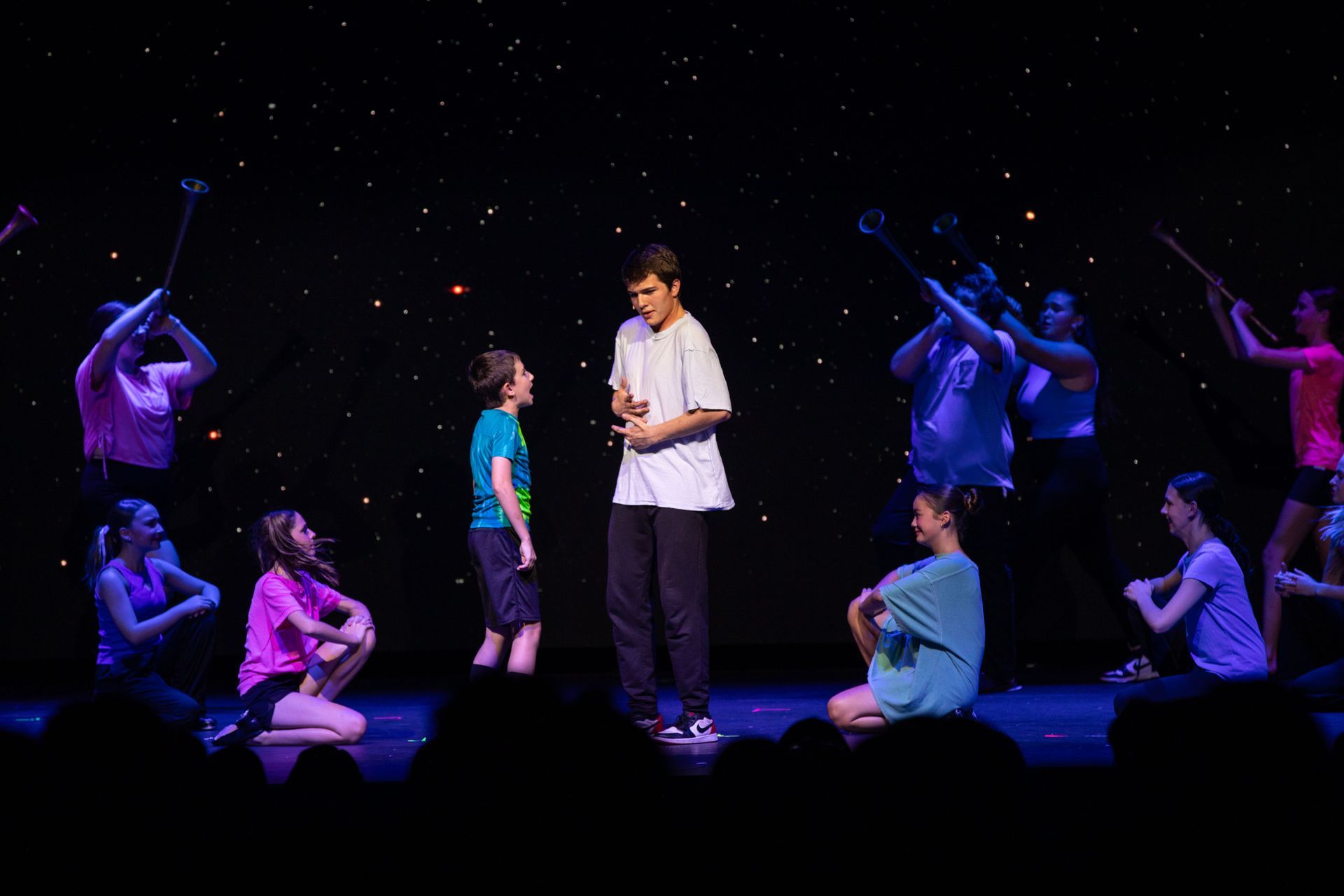 Actors on a dark stage under purple lights, surrounded by seated performers in a theatrical scene