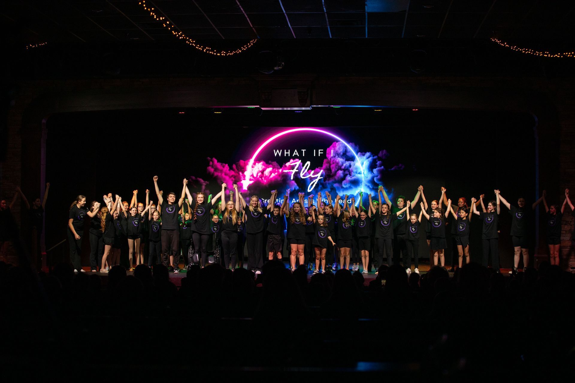 Large group on stage at a concert, raising arms before a bright pink and blue circular light display