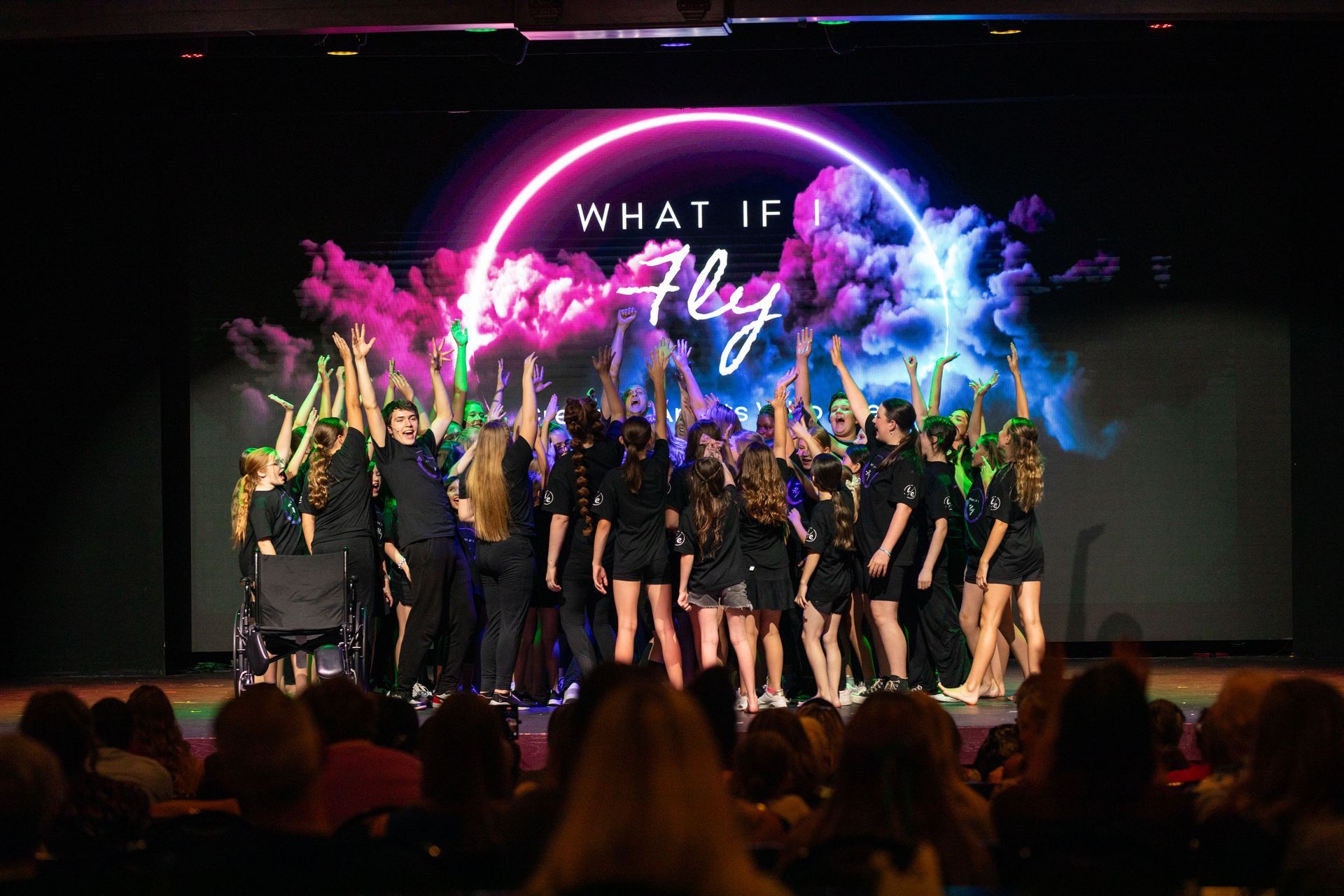 Dance troupe performing on stage with pink and blue smoke under a “What If” backdrop