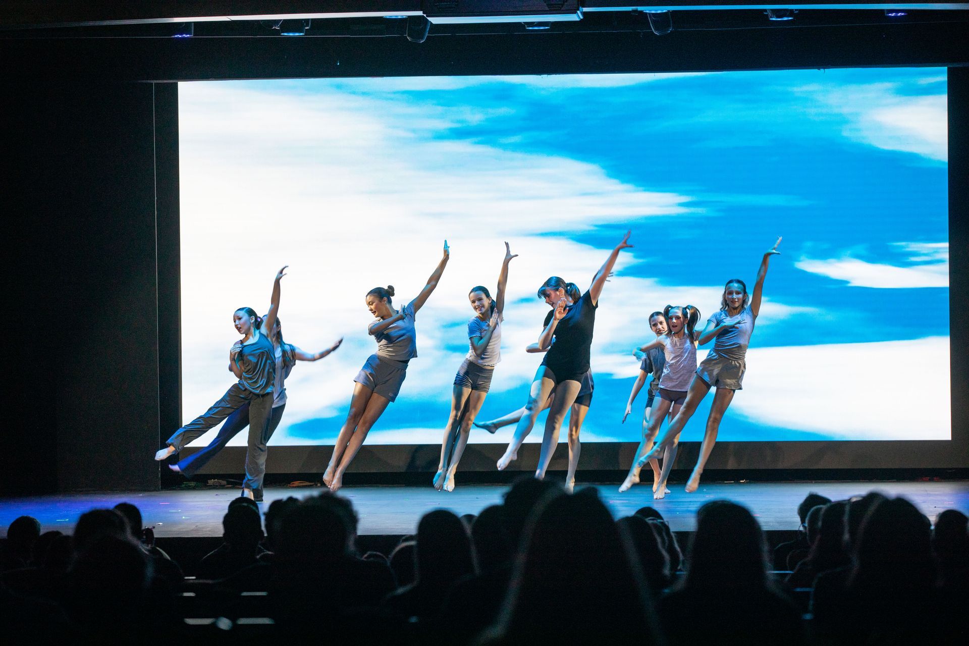 Five dancers in blue costumes performing on stage under bright lights with a large screen backdrop