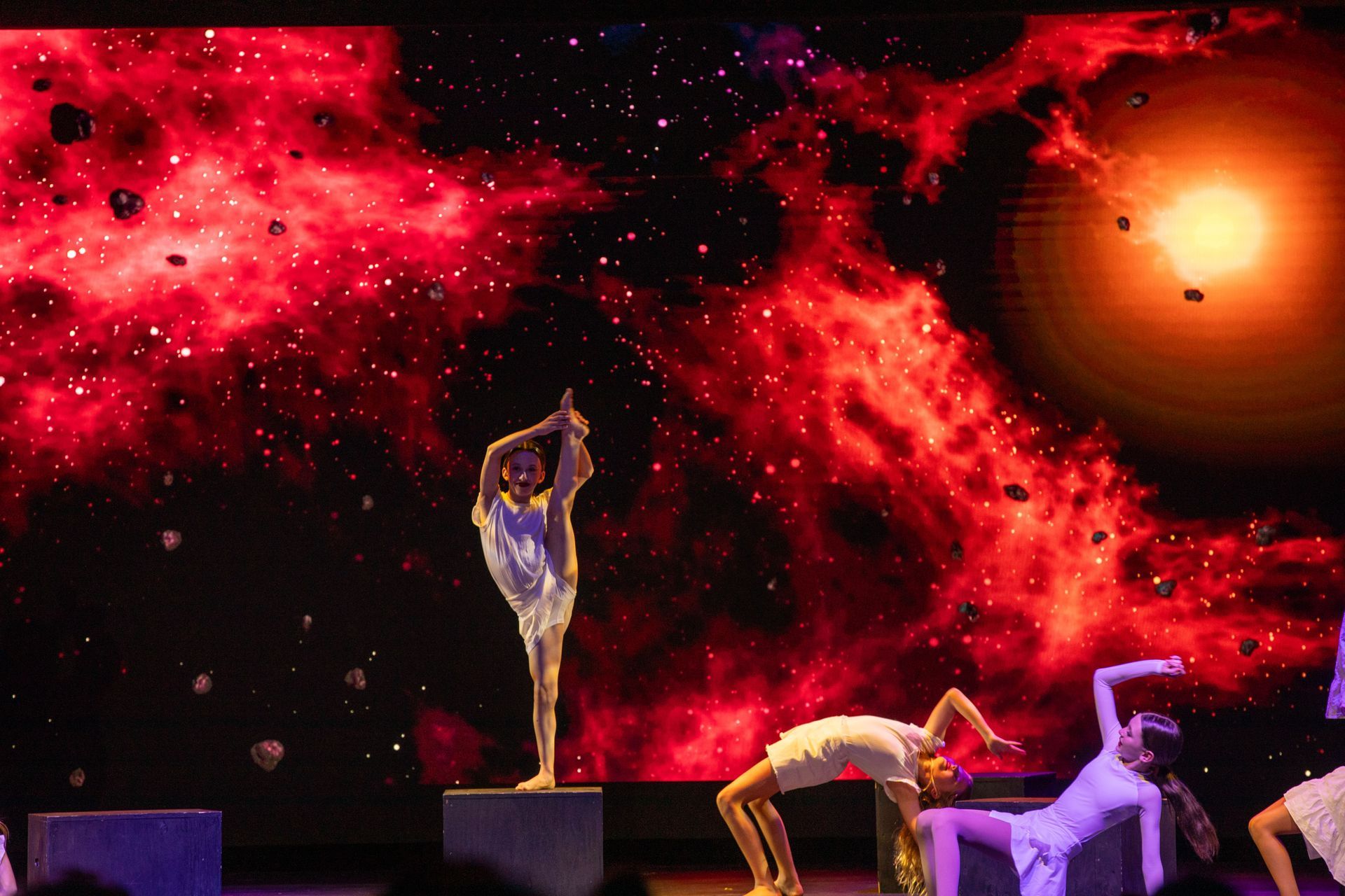 Dancers on stage under red lighting with a fiery orange backdrop and one performer balanced in a pose