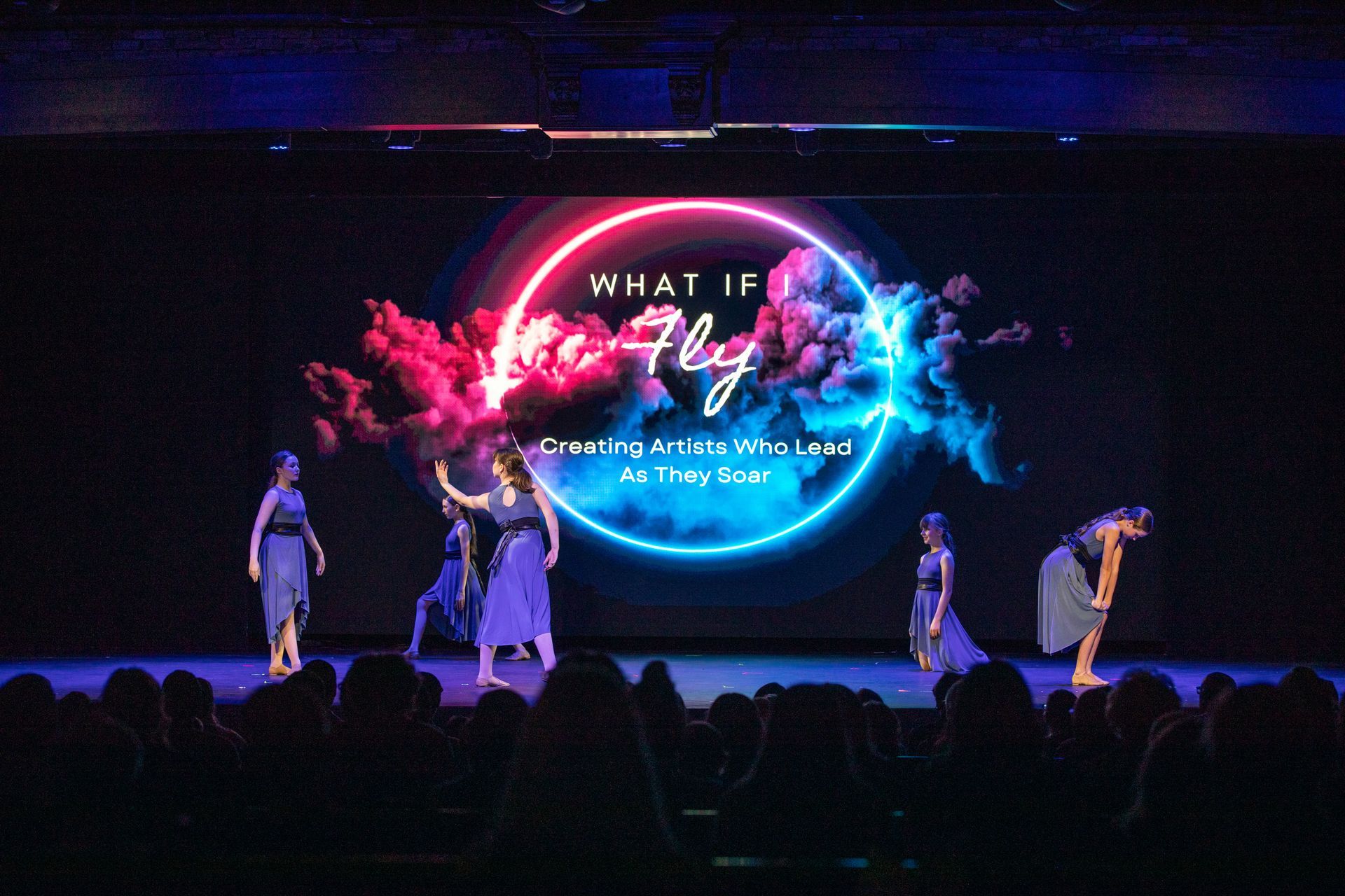 Dancers on a stage before a neon blue and pink circular backdrop with text and crowd silhouettes