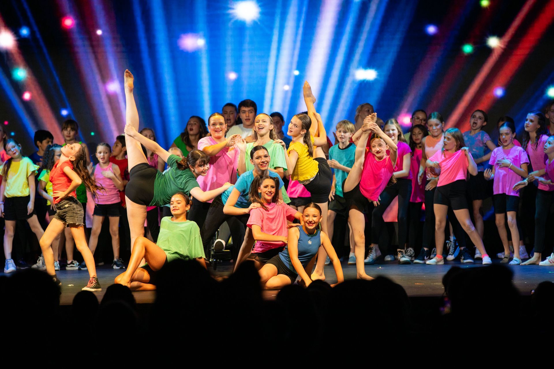 Large group of dancers on stage under colorful spotlights, posing and performing in bright costumes