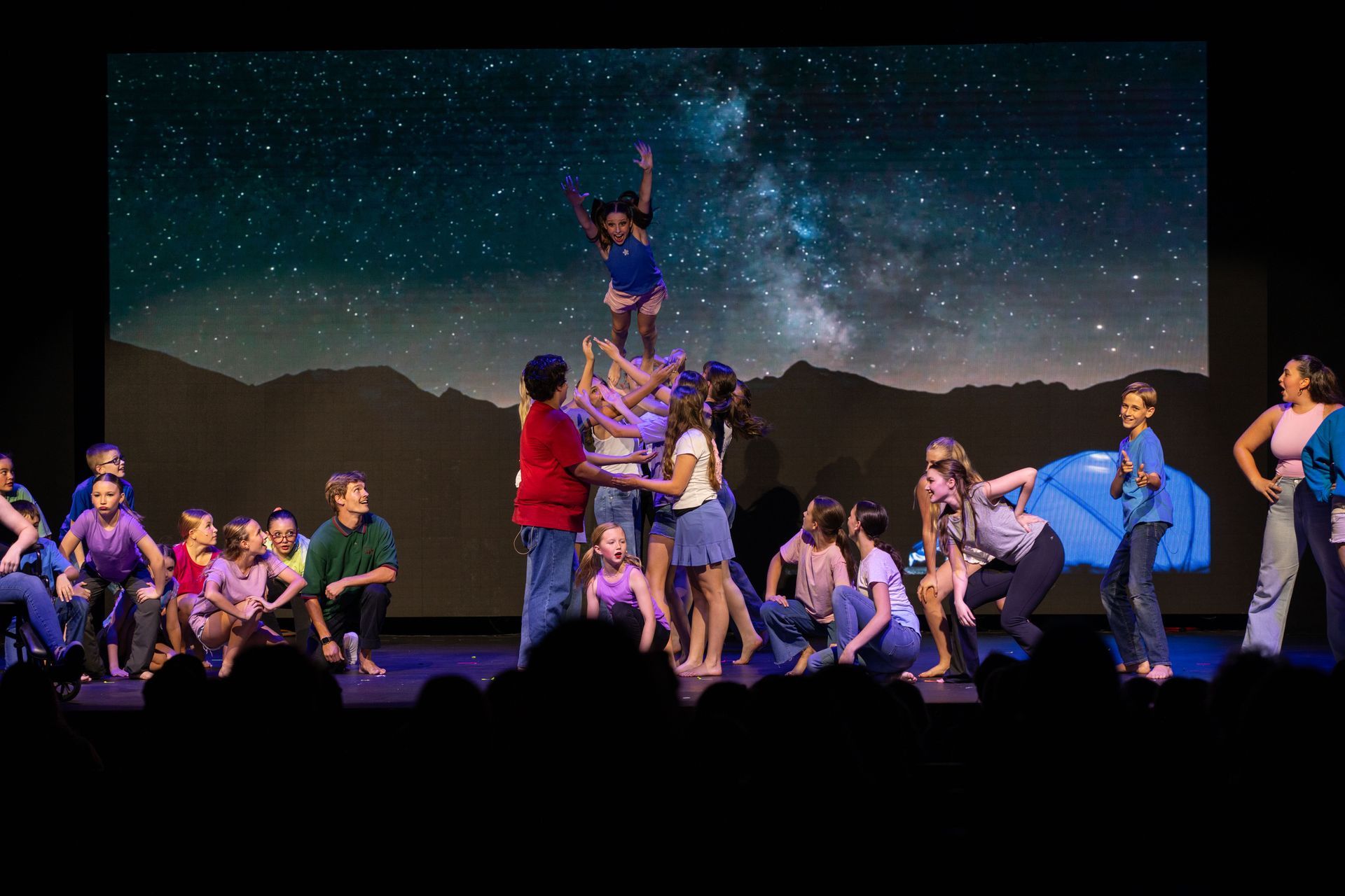 Dance troupe performing on stage under a starry backdrop, with one dancer lifted center stage.
