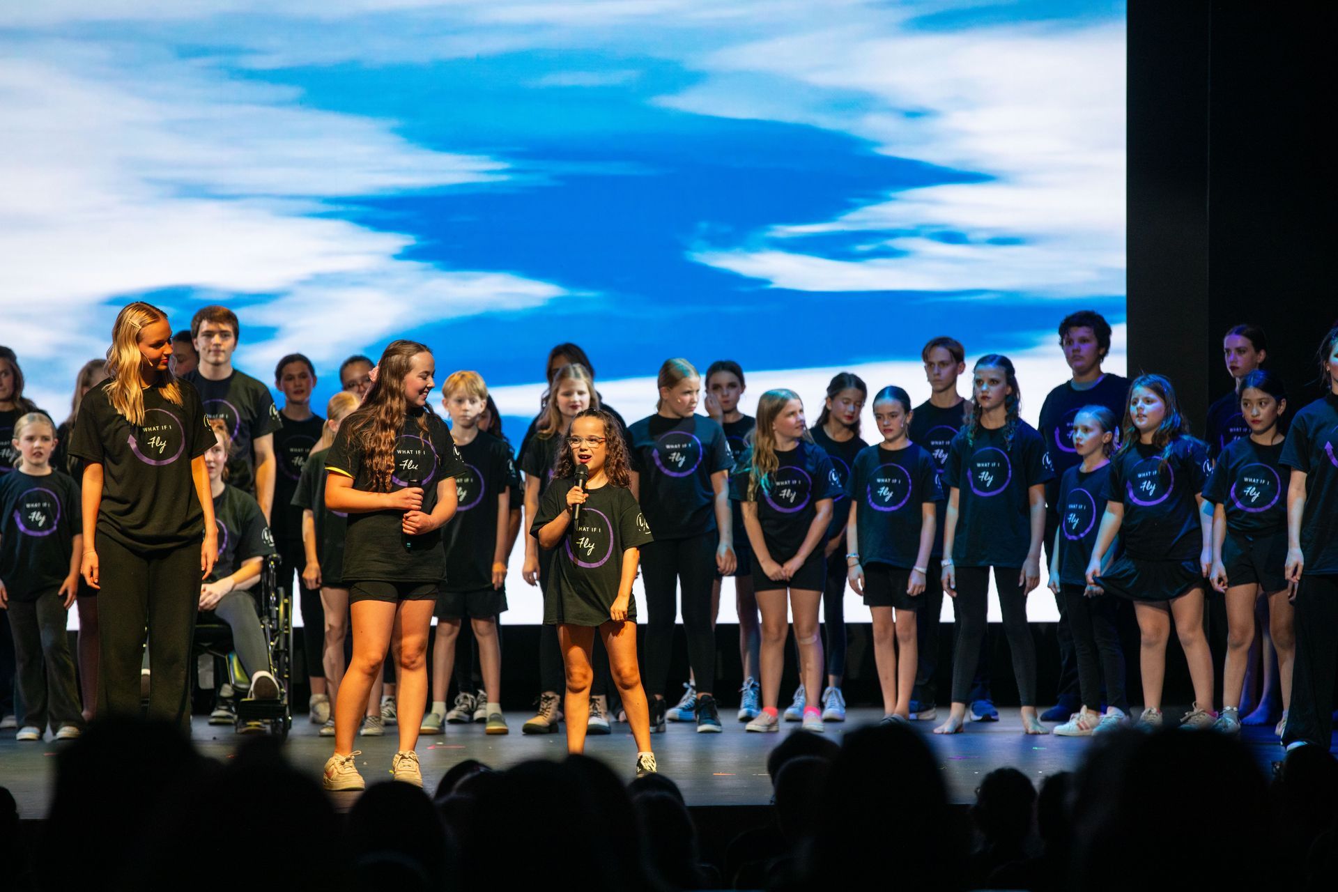 Group of performers in black and blue outfits standing on a stage under a bright sky backdrop