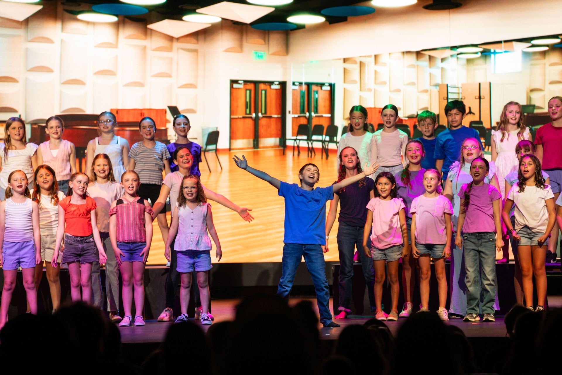 Children singing on a stage, led by a conductor, in a brightly lit auditorium