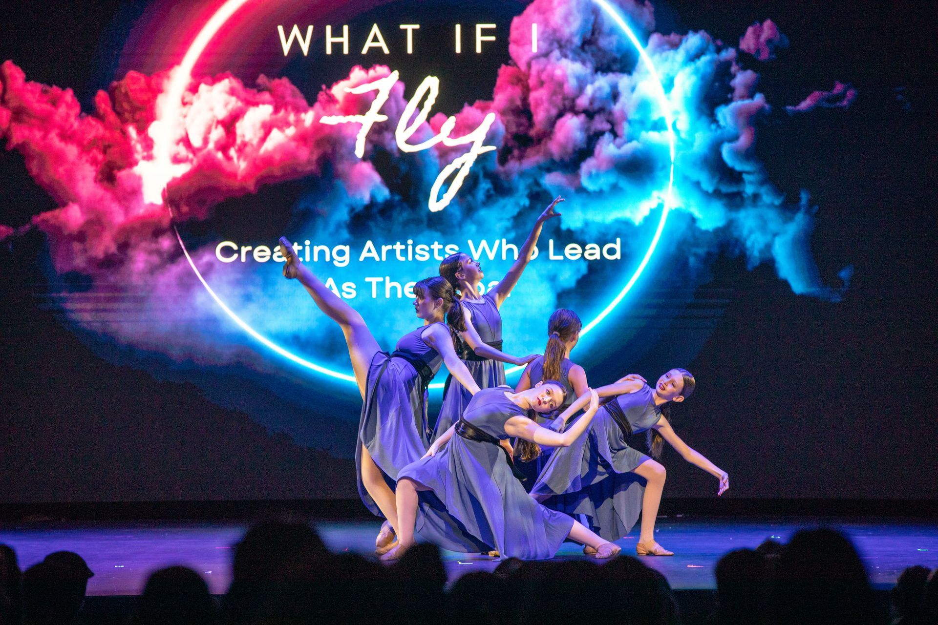 Dancers onstage beneath a neon pink and blue “What If Fly” backdrop with “Creating Artists...” text