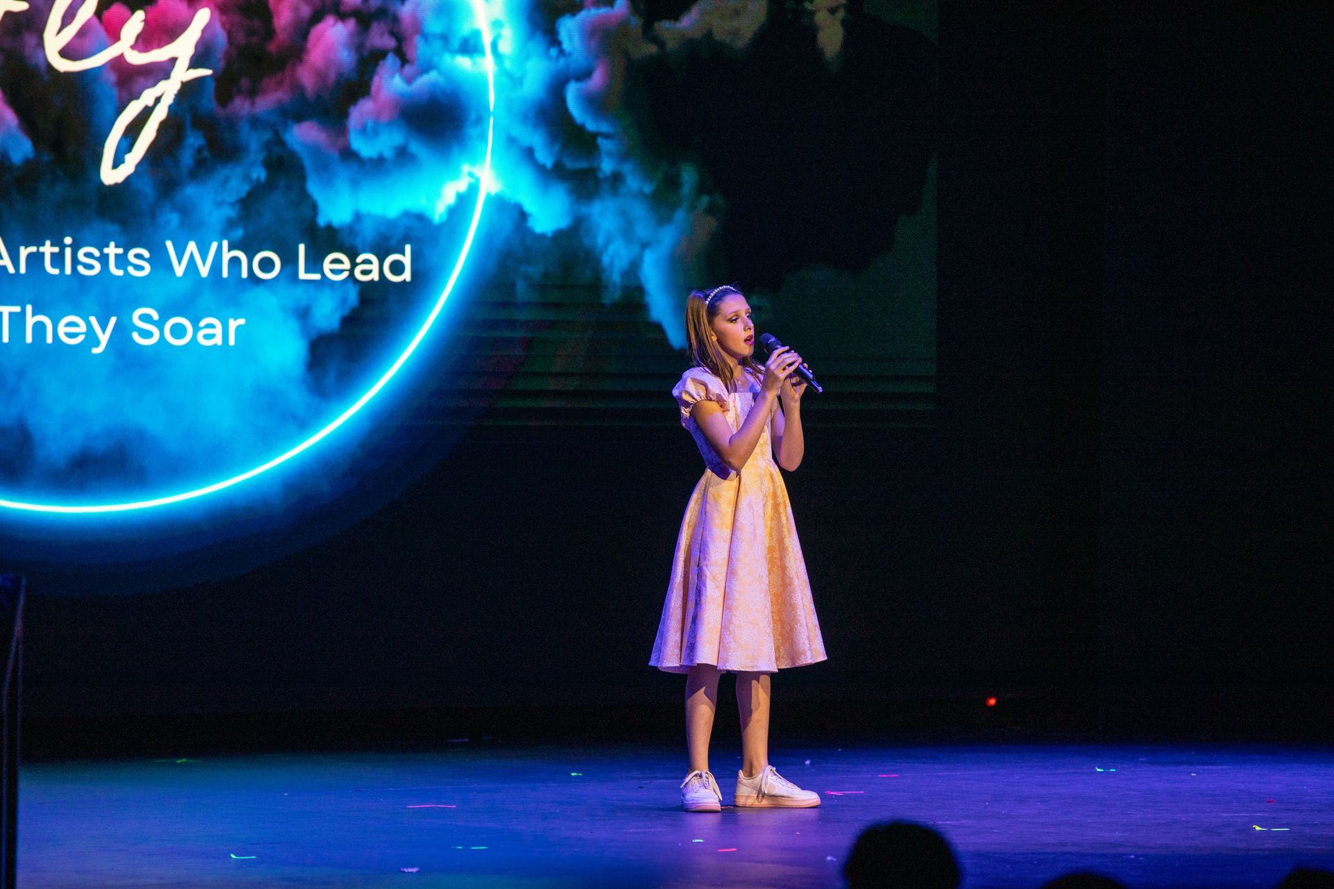 Young singer in a white dress performing on stage beside text reading “Artists Who Lead, They Soar”