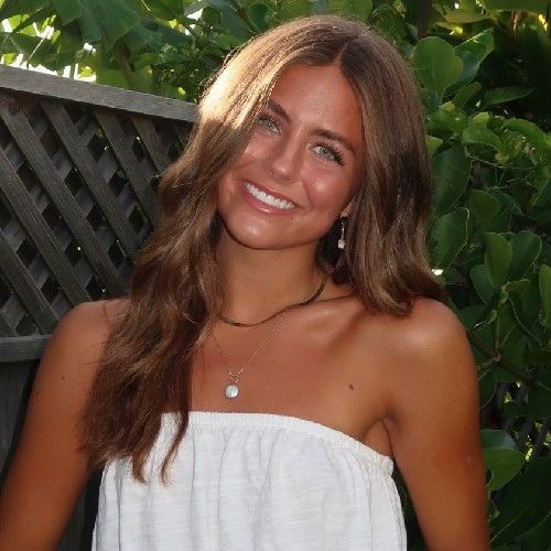 Smiling woman in a white strapless top standing outdoors by a leafy fence