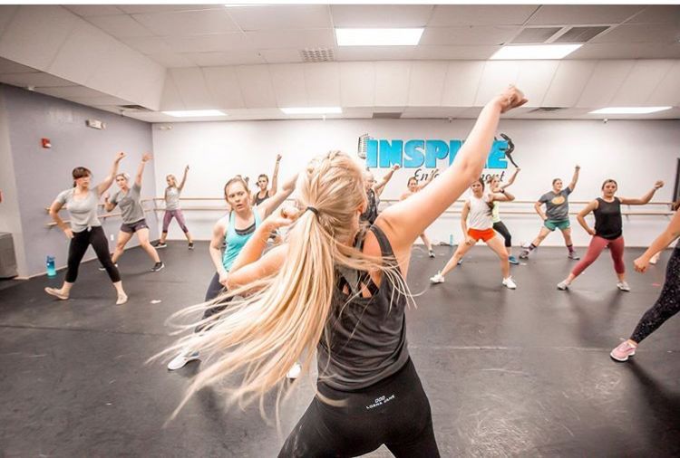 Group fitness class in a studio, instructor leading dancers with arms raised in bright workout clothes.