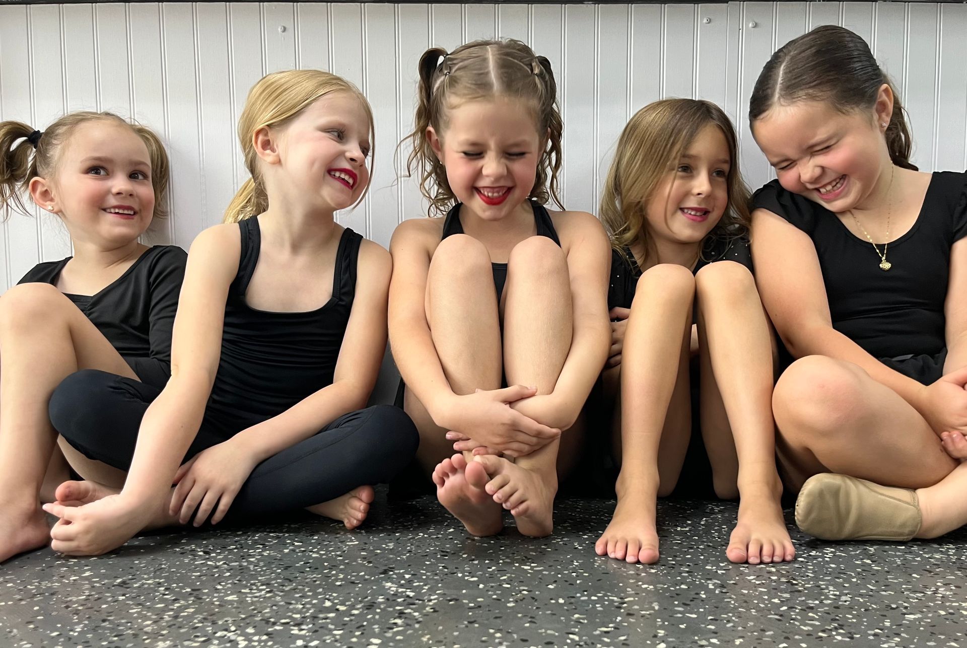 Five children in black outfits sitting on a floor, smiling and posing together