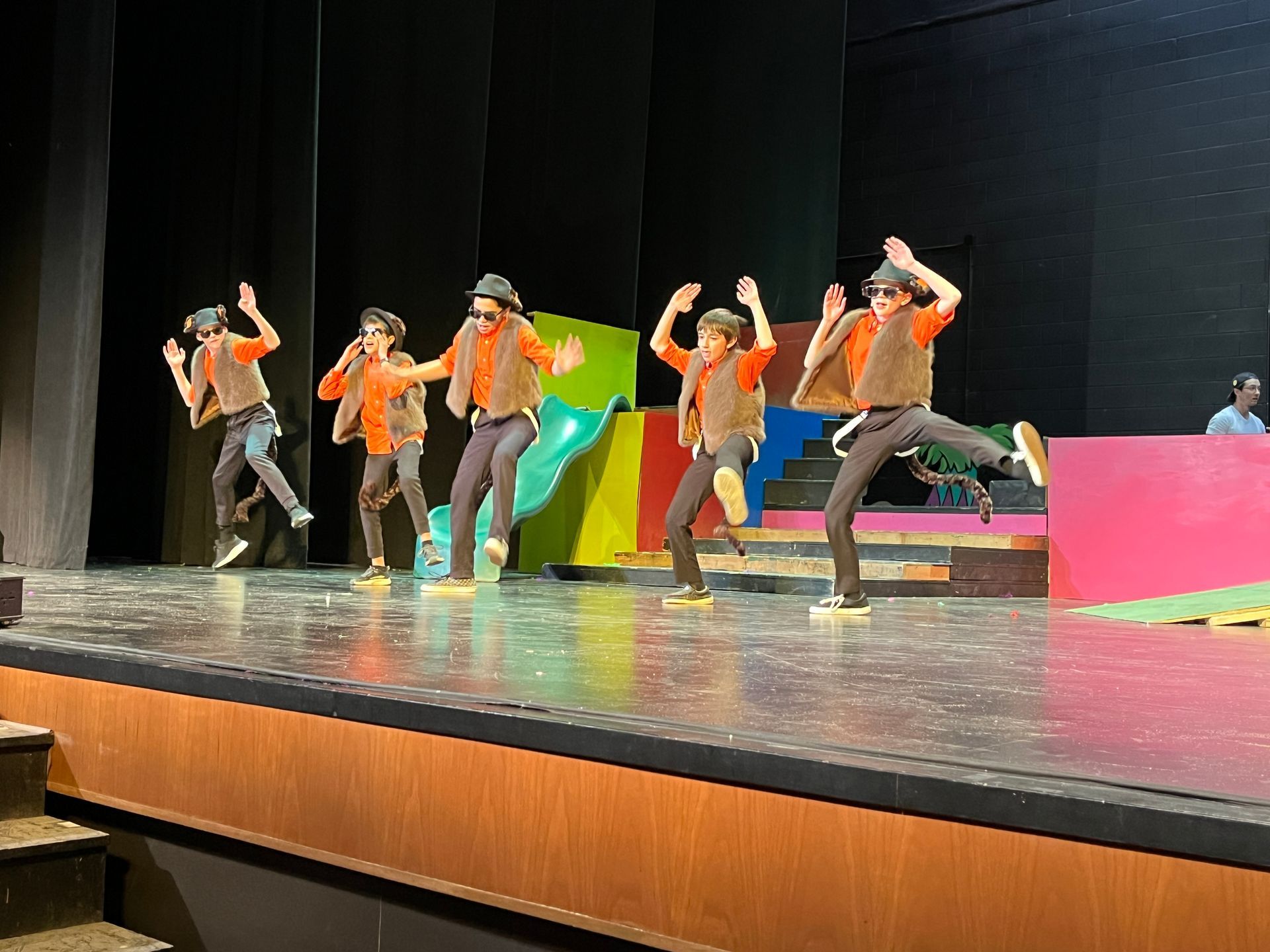 Five dancers in orange jackets perform on a stage with colorful props and a black backdrop.
