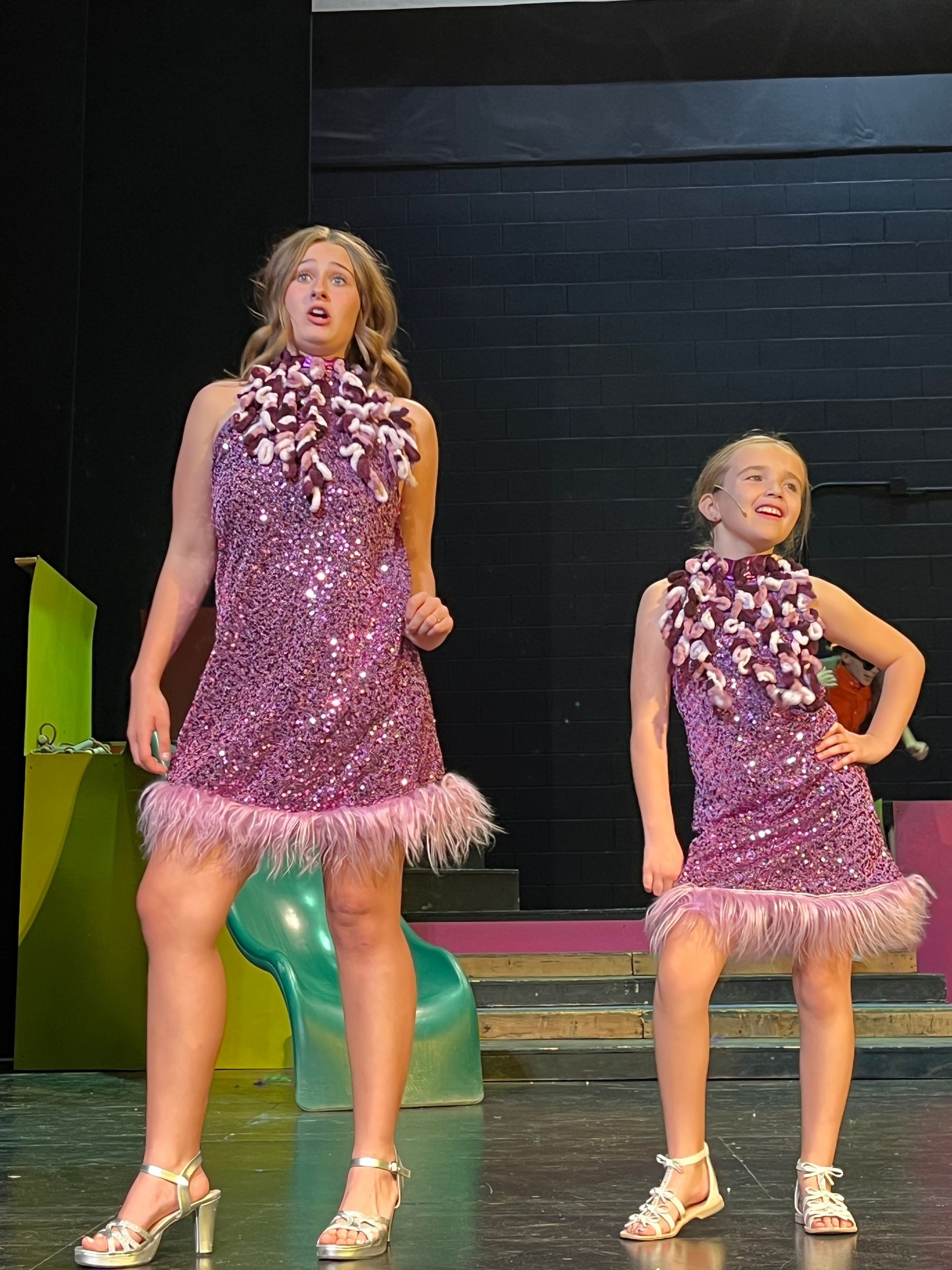 Two dancers in matching purple fringe costumes pose onstage against a dark backdrop.
