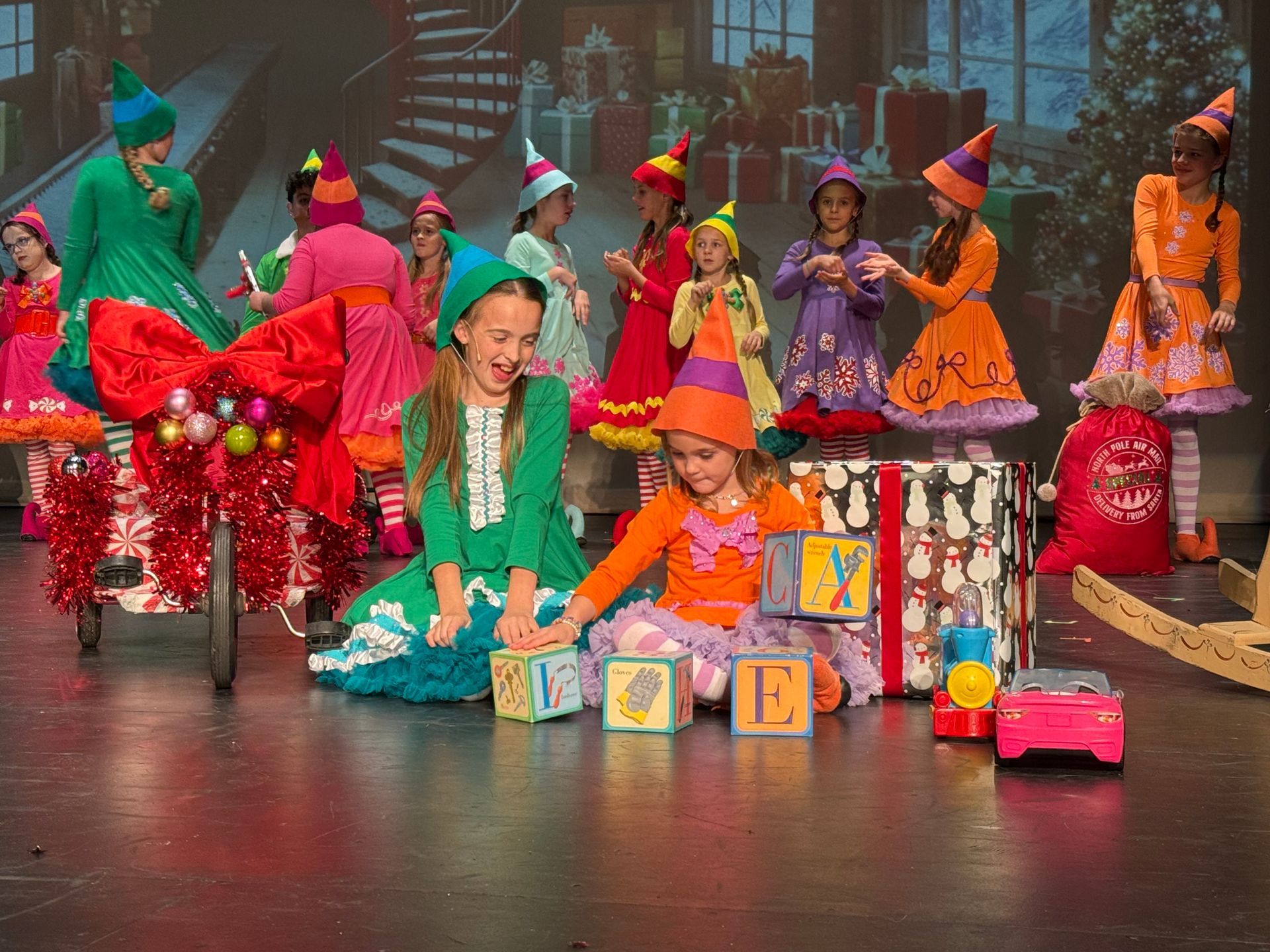 Children in colorful costumes sit and stand with toy blocks on a stage, suggesting a festive performance
