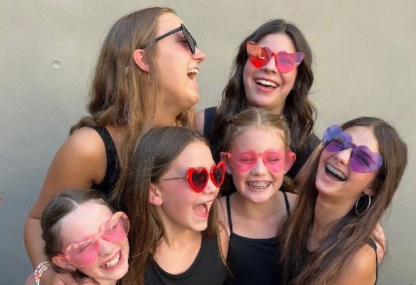 Six smiling friends wearing colorful heart-shaped sunglasses, posing together outdoors.