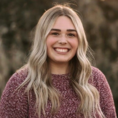 Smiling person with long blonde hair wearing a maroon sweater outdoors against a blurred background