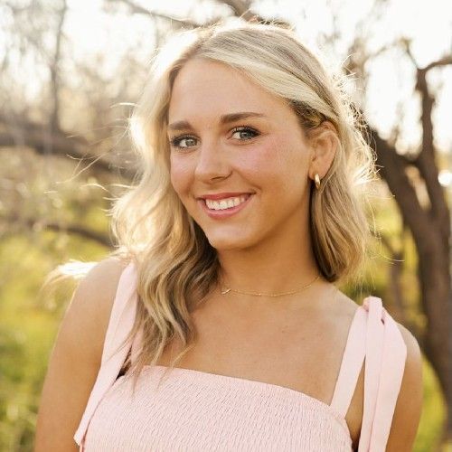 Smiling woman in a light pink dress outdoors with soft sunlight and trees in the background