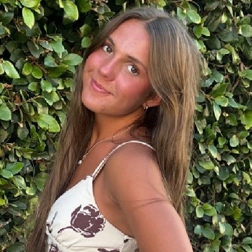 Woman smiling in a white floral dress, standing before a leafy green hedge.