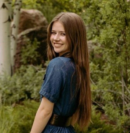 Smiling woman in a blue dress standing in a lush green garden, looking over her shoulder.