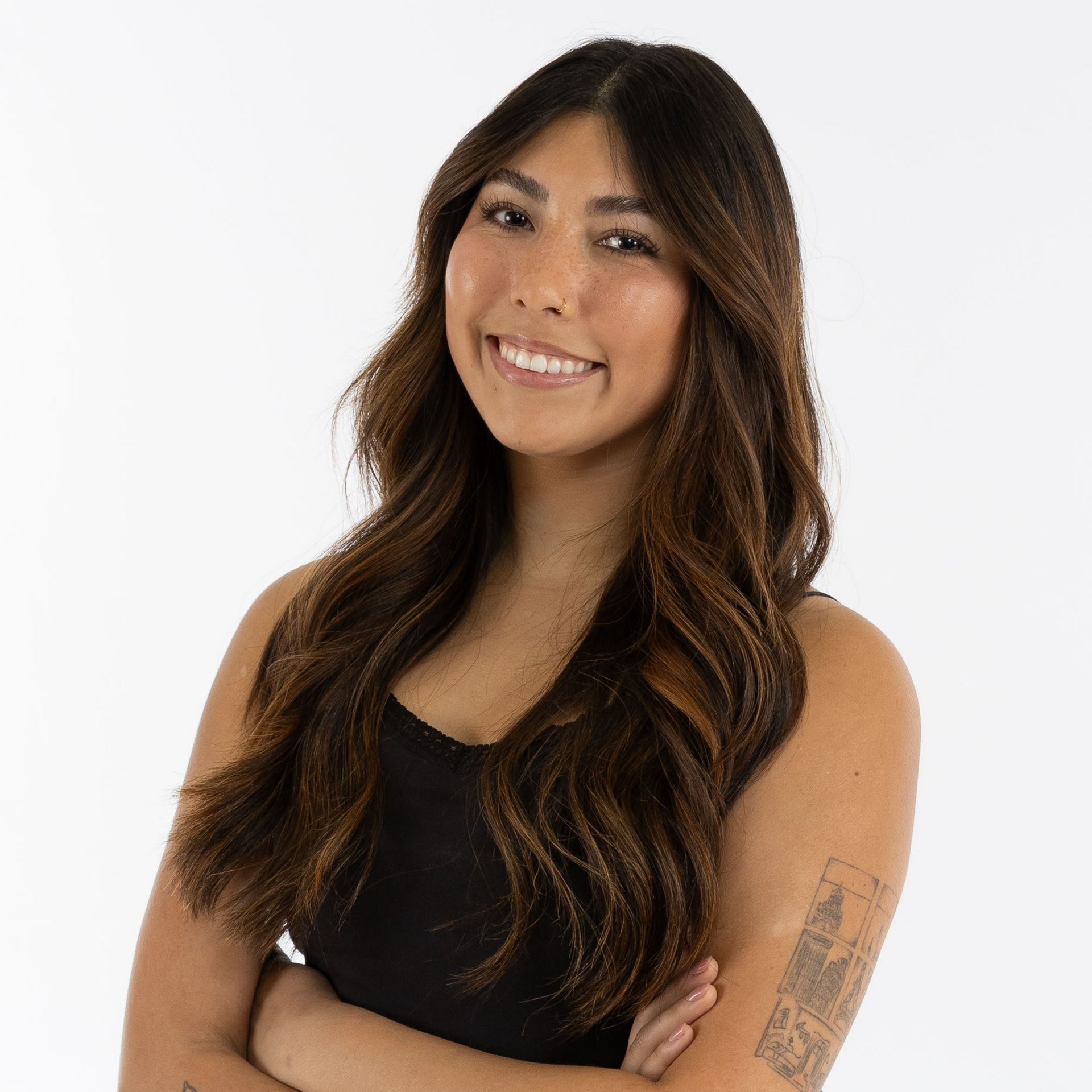 Smiling woman with long wavy brown hair in a black sleeveless top, standing with arms crossed on a white background