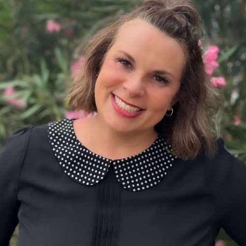 Smiling woman in a black top with a white polka-dot collar, standing outdoors with pink flowers in the background