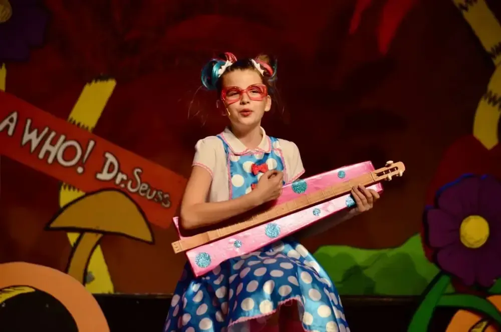 Person in pink glasses and blue polka-dot dress holding a pink toy guitar on a colorful Dr. Seuss stage