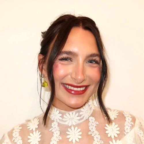 Smiling woman with dark hair, red lipstick, and a white floral lace top against a plain background