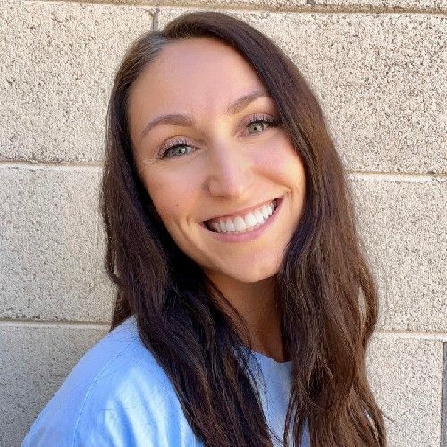 Smiling woman with long brown hair standing against a light stone wall, wearing a light blue shirt