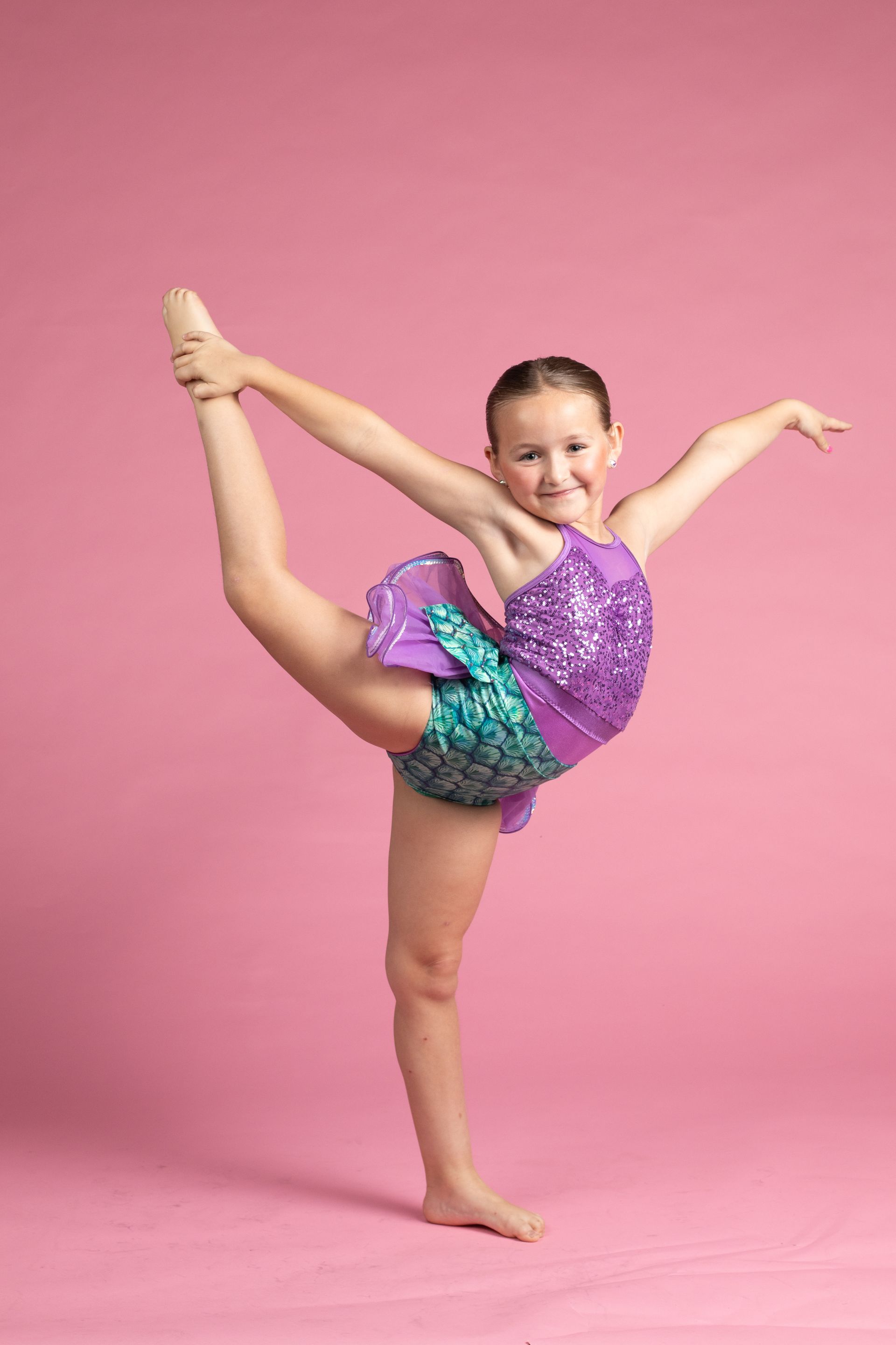 Young gymnast posing on one leg against a pink studio backdrop, wearing a purple and turquoise leotard