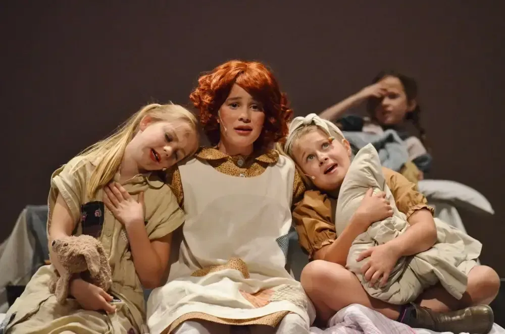 Three children sit onstage, leaning together in costumes, with a woman seated behind them.