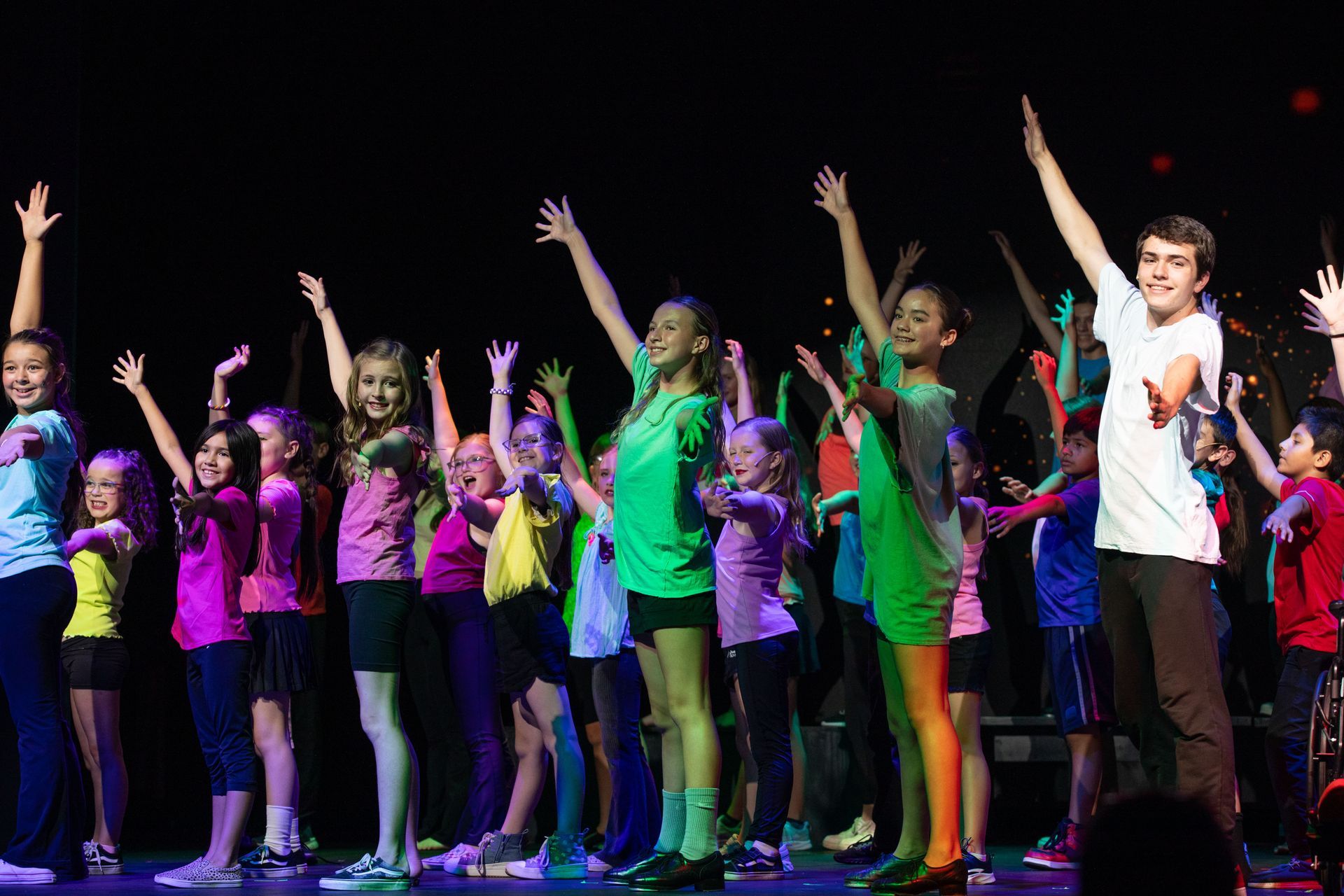 A group of dancers in colorful outfits perform on stage with arms raised under bright lights.