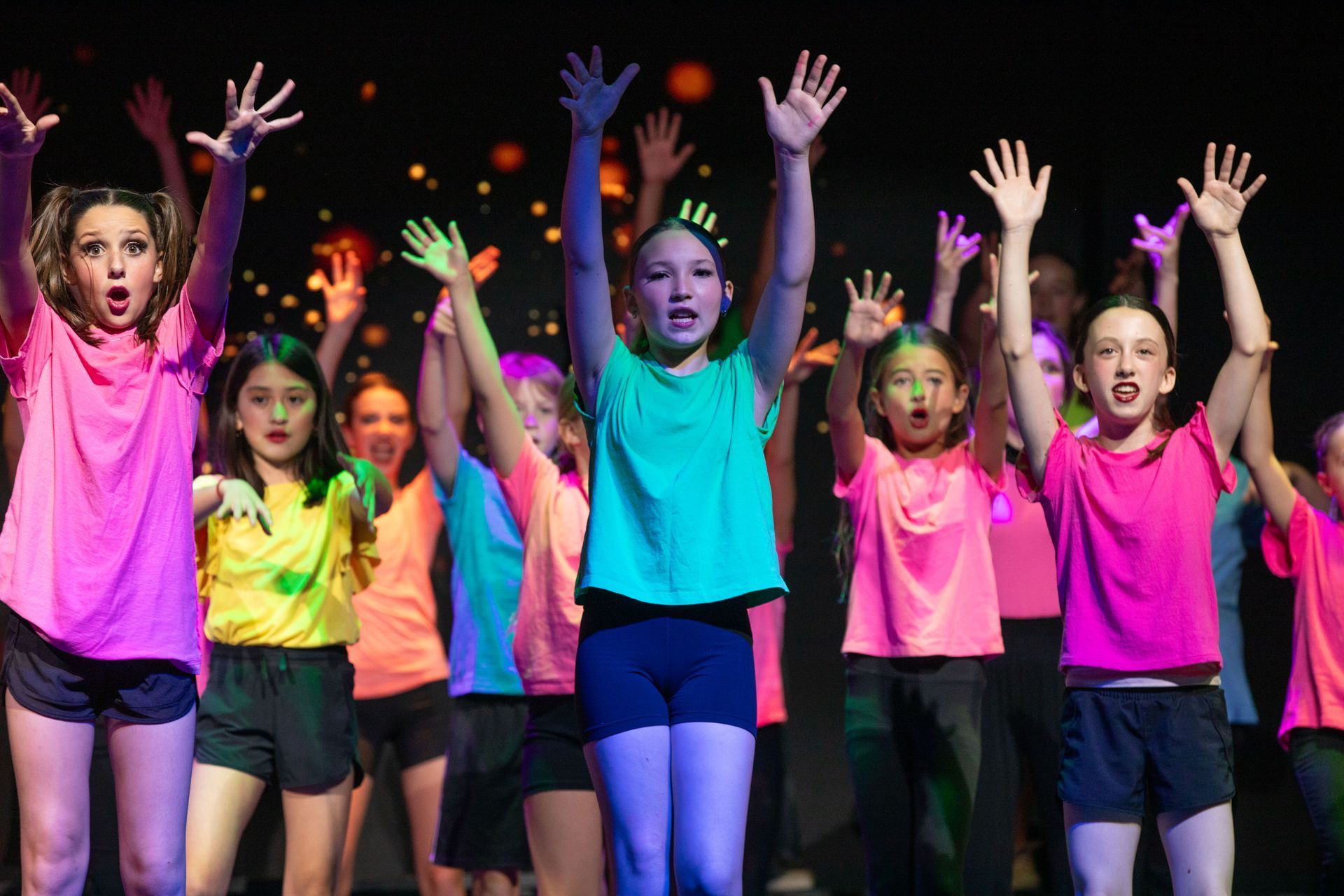 Children in colorful shirts dance onstage with arms raised under bright stage lights