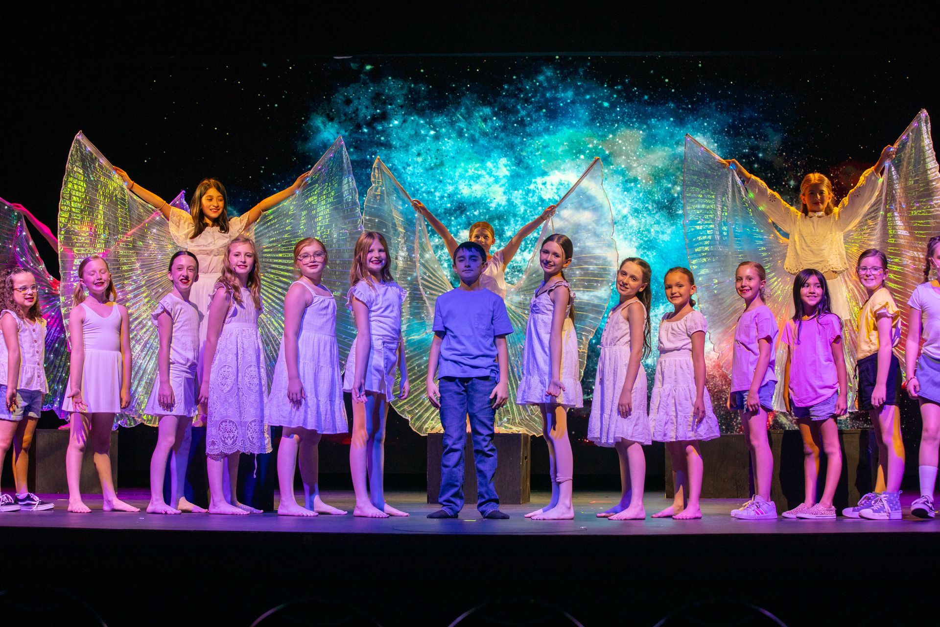 Children in white costumes standing on a stage under colorful blue and purple lights