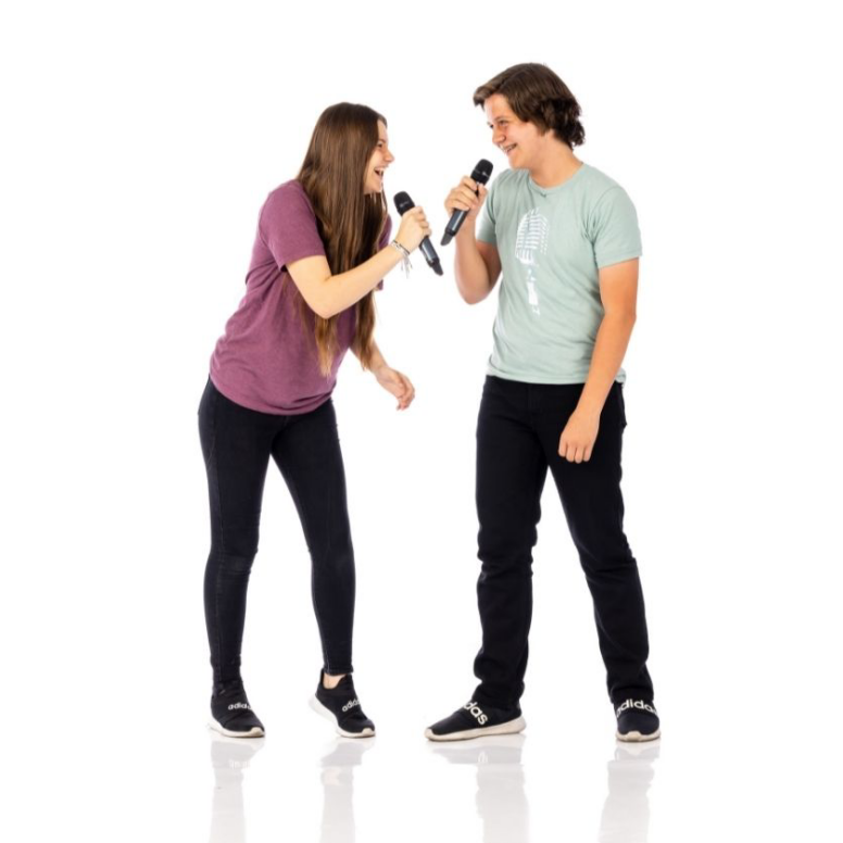 Two people singing into microphones, facing each other on a white background.
