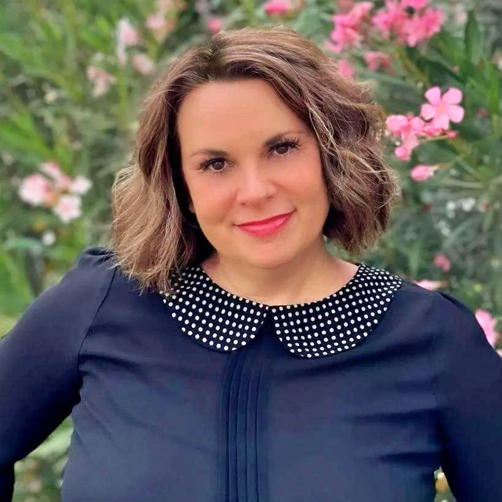 Portrait of a smiling woman in a navy blouse with a polka-dot collar, standing among pink flowers.