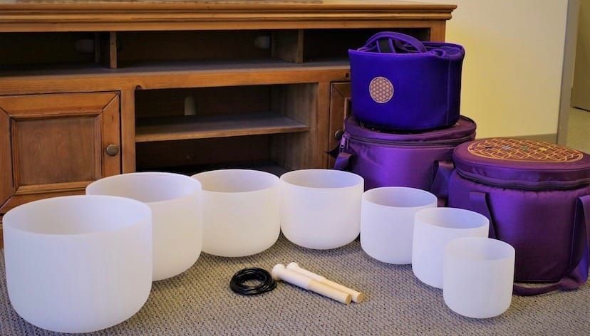 White singing bowls and purple carrying cases arranged on a carpeted floor in front of a wooden cabinet