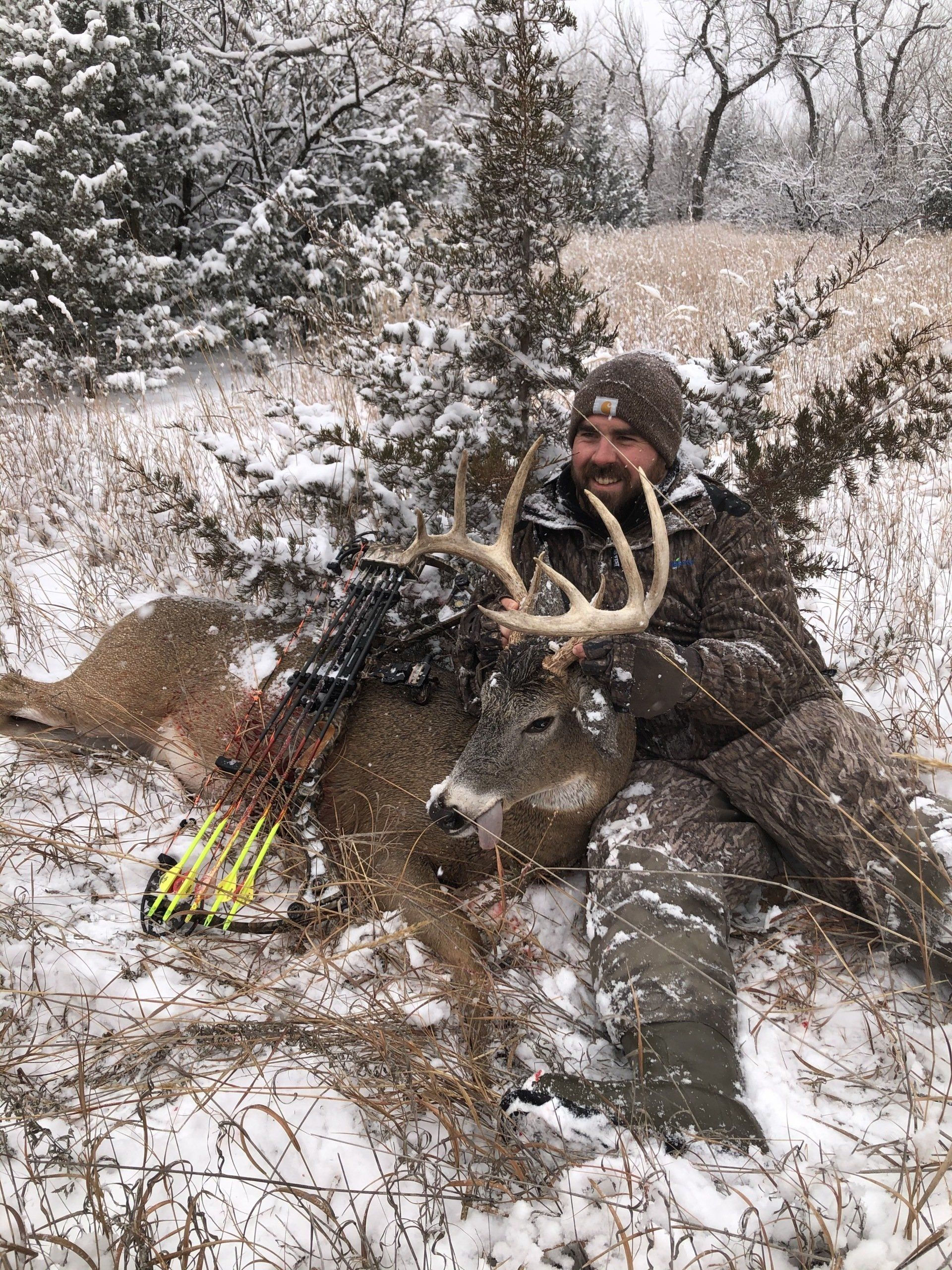 Hunter kneeling in snowy woods beside a large deer with antlers and a bow nearby