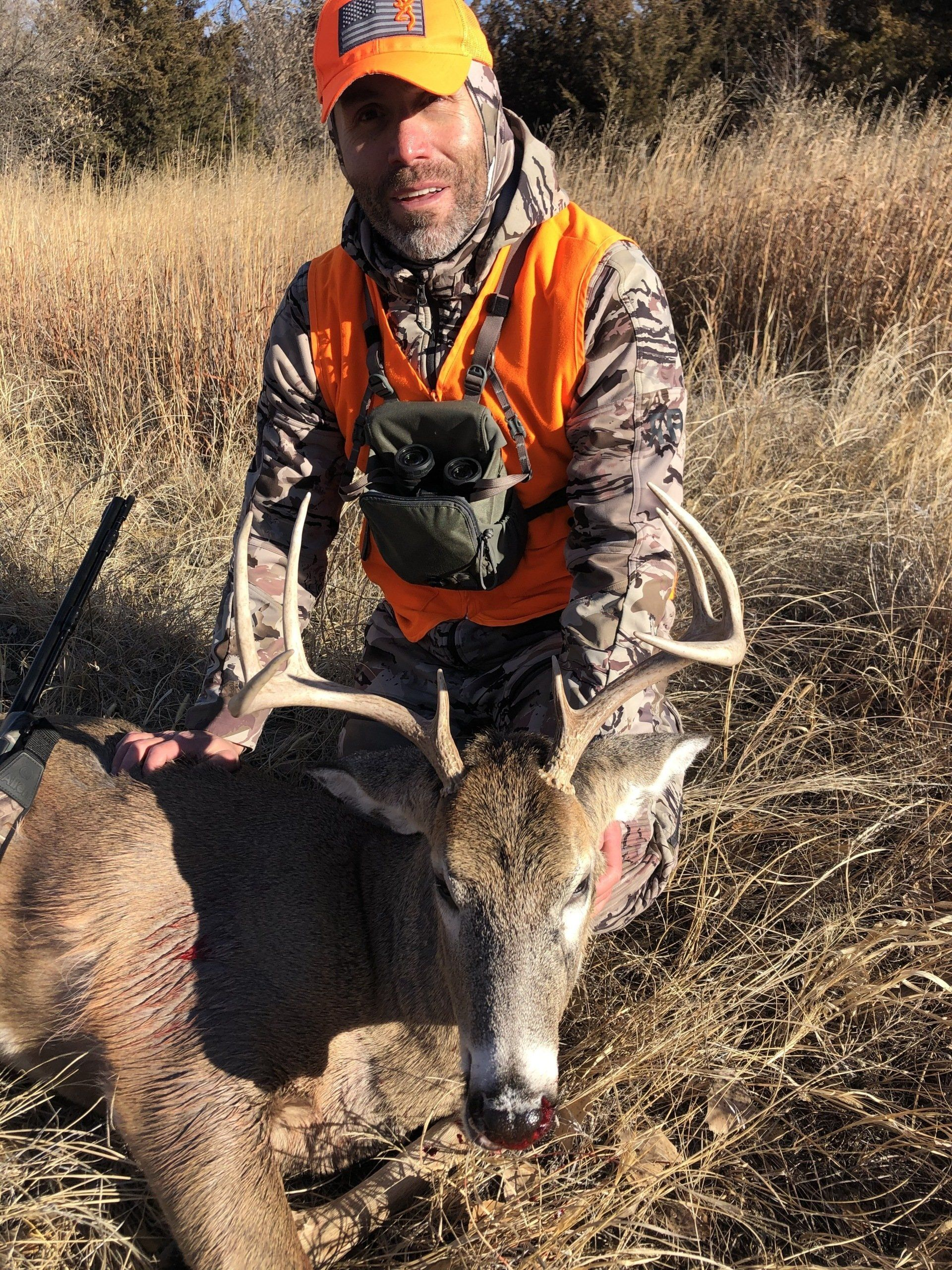 Hunter in orange vest kneeling beside a deer in tall dry grass