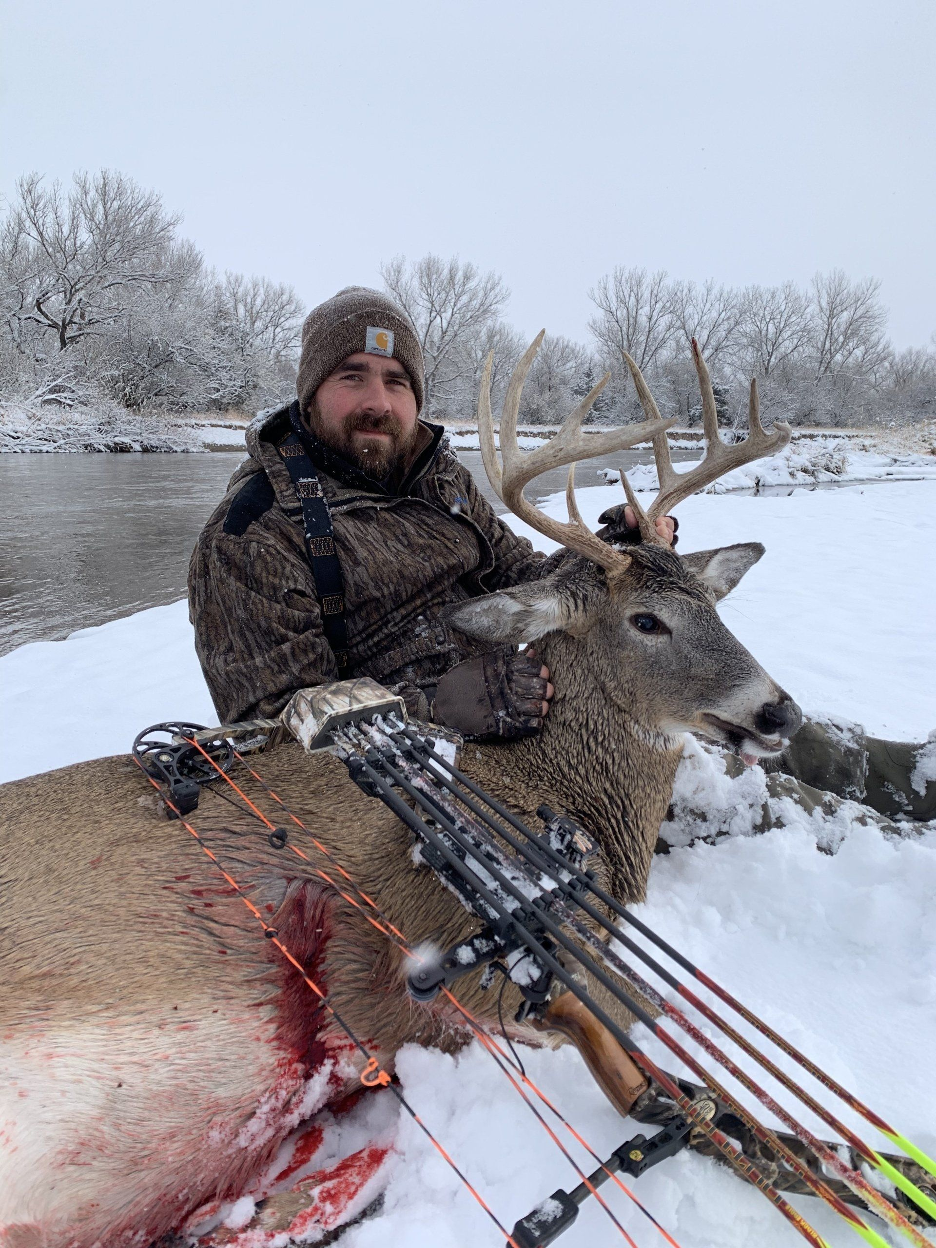 Hunter kneels beside a deer in a snowy field, holding a crossbow, with frosty trees in the background