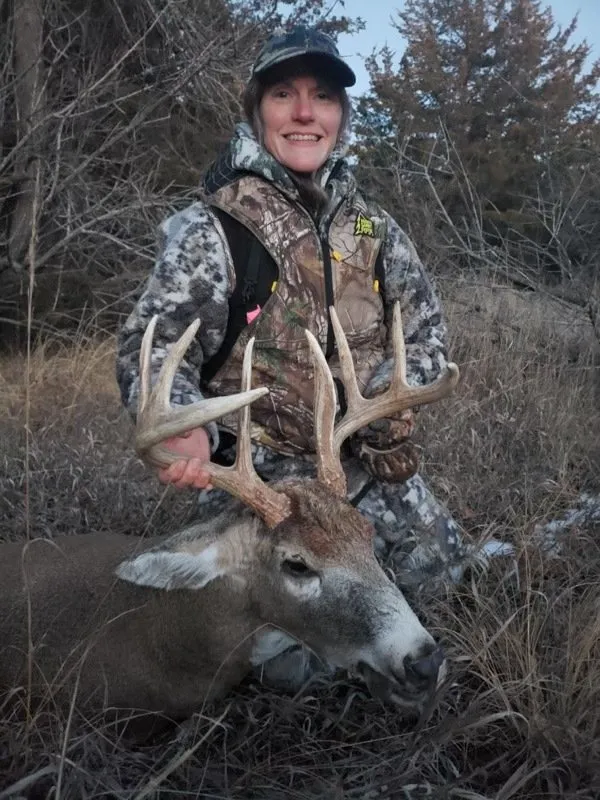 Hunter posing with a large deer in a wooded area at dusk, wearing camouflage and a cap.