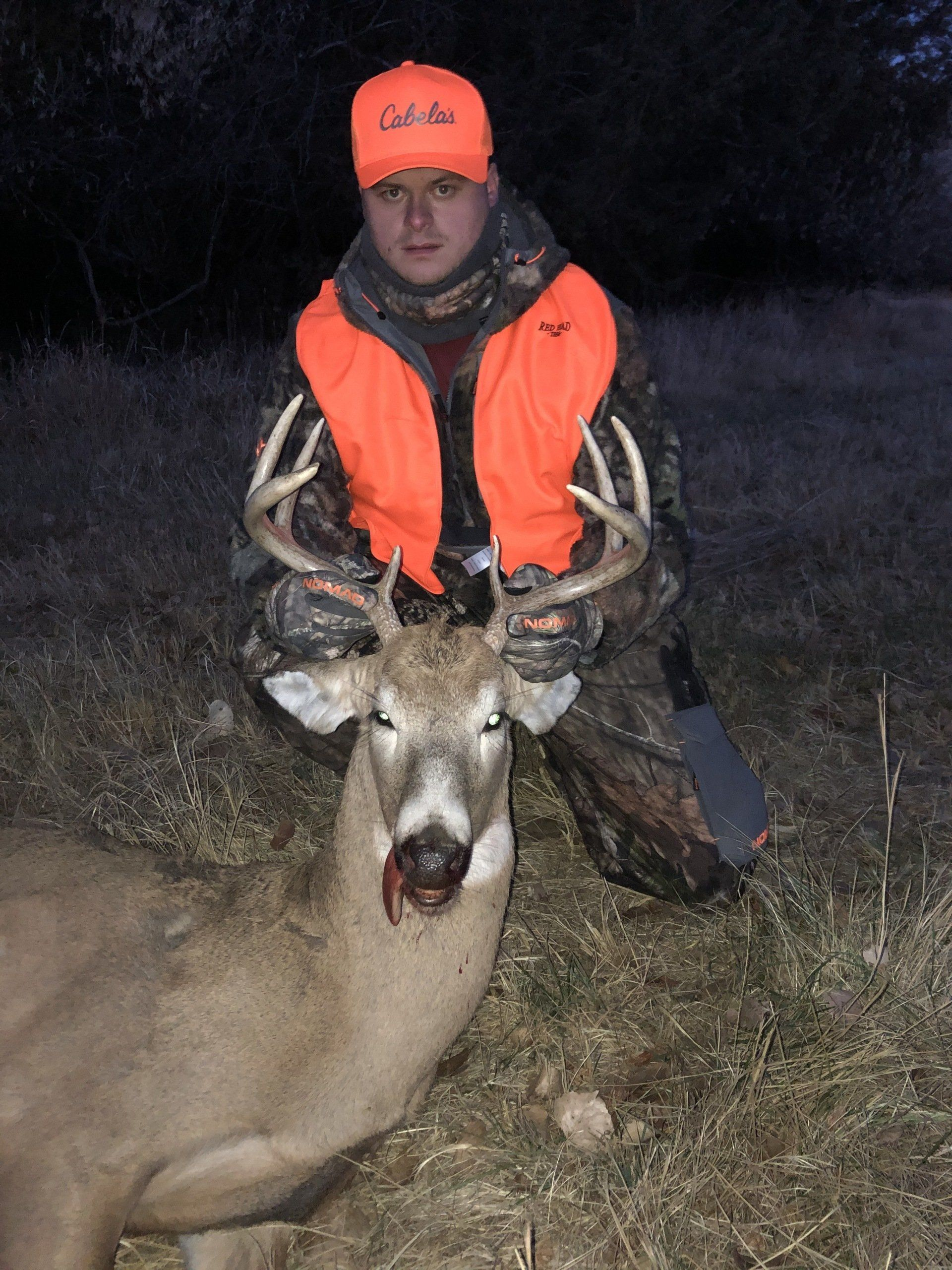 Hunter in orange vest and cap kneels beside a large buck at night.