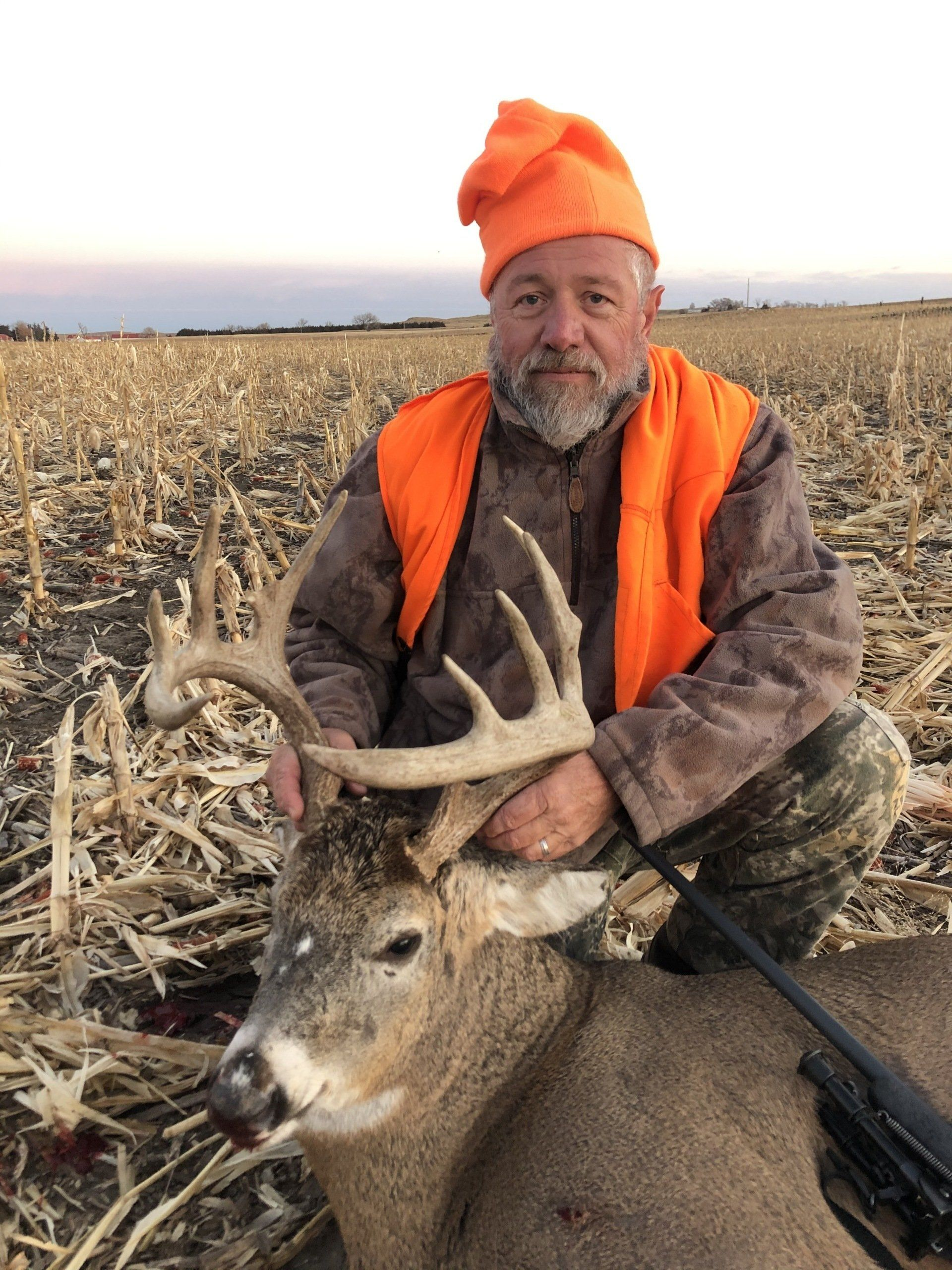 Hunter in orange vest and hat posing with a deer in a harvested field at sunset